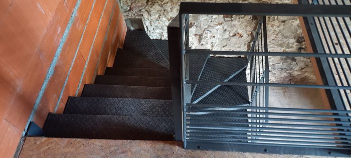 Escalier intérieur tournant à marches en tôle larmée et garde-corps à barreaudage, contre un mur en pierre