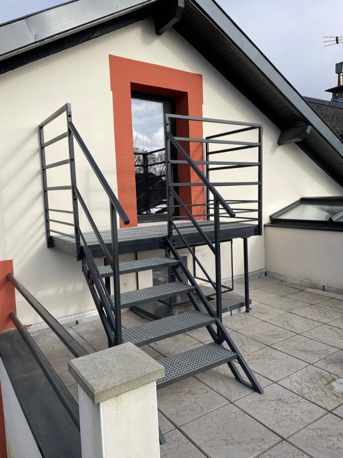 Escalier extérieur anthracite à marches caillebotis et palier desservant une porte-fenêtre à l'étage, sur une terrasse
