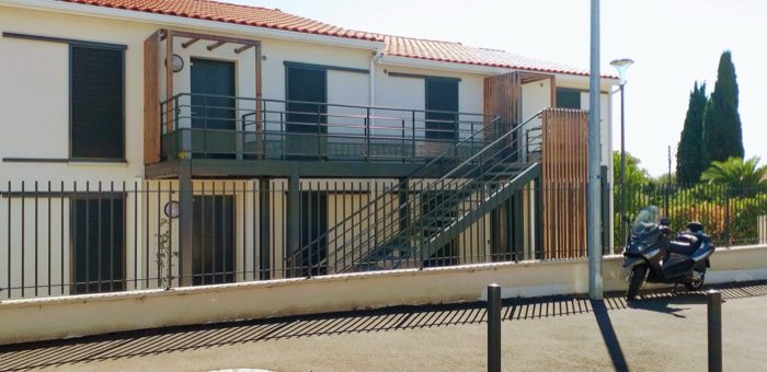 Garde-corps et escalier extérieurs noirs bordant une terrasse sur pilotis, le long d'un bâtiment
