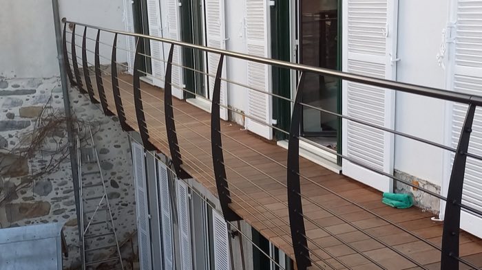 Garde-corps à montants galbés et lisses horizontales bordant une coursive en bois, contre un mur en pierre