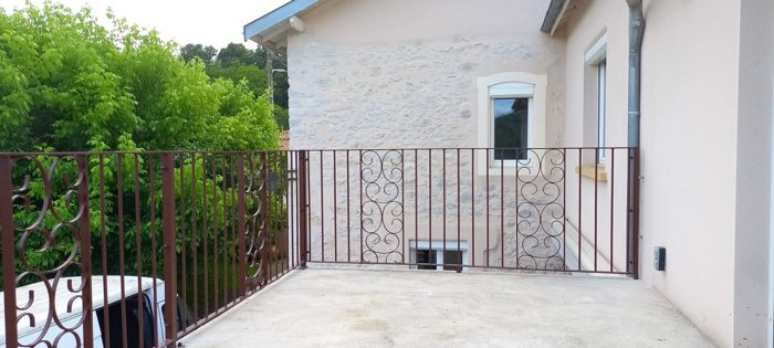 Garde-corps de terrasse en fer forgé brun à panneaux de volutes, devant une façade en pierre