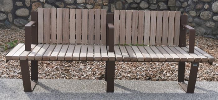 Double banc public à lattes de bois et structure acier brun, devant un mur en galets