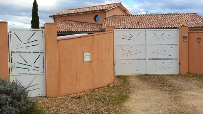 Portail battant blanc à panneaux pleins ajourés de fentes obliques, avec portillon assorti, devant une maison à façade ocre