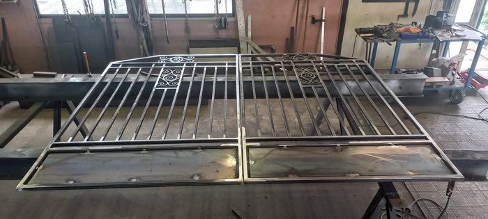 Deux vantaux de portail en acier brut posés à plat dans l'atelier avant traitement, barreaudage et volutes décoratives