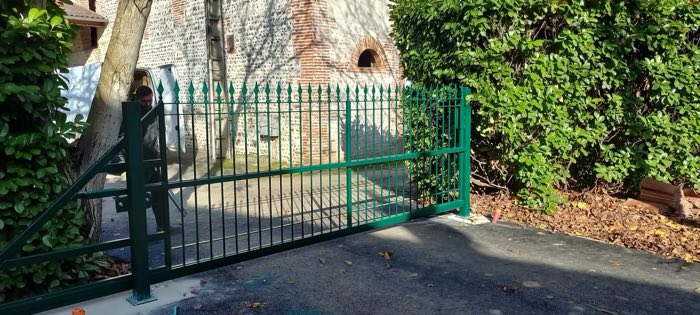 Portail coulissant vert à barreaudage et pointes de lance, devant une bâtisse en brique et galets