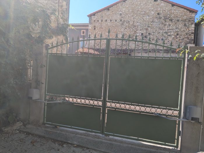 Portail battant vert en fer forgé, motorisé, à panneaux pleins et frise d'anneaux, dans une rue de village